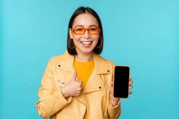 Jonge, stijlvolle chinese vrouw die het scherm van de mobiele telefoon laat zien en een smartphone-app laat zien die over een blauwe achtergrondkopieerruimte staat