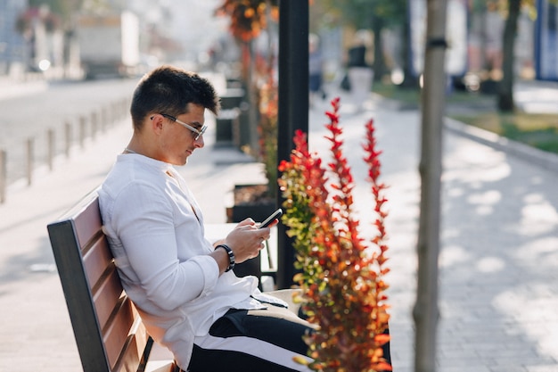 Jonge stijlvolle man in shirt met telefoon op bankje op zonnige warme dag buitenshuis