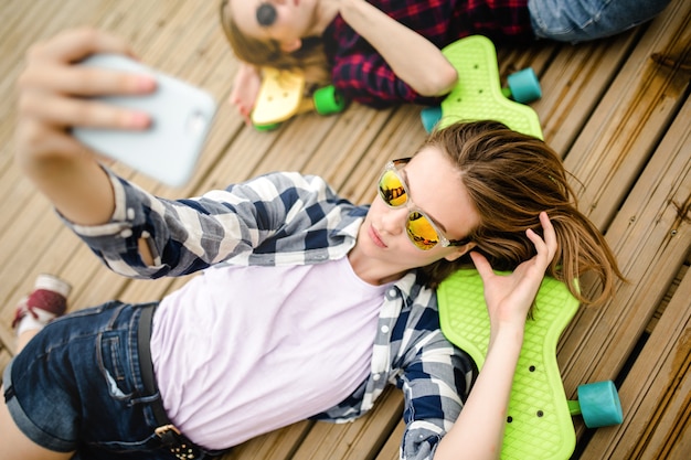 Jonge stijlvolle stedelijke meisje in hipster outfit selfie maken terwijl liggend met op houten pier.