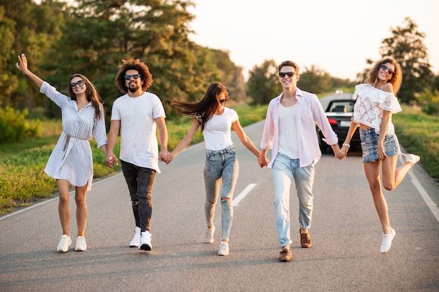 Jonge stijlvolle twee jongens en drie meisjes houden elkaars hand vast en lopen op een zonnige dag op de weg.