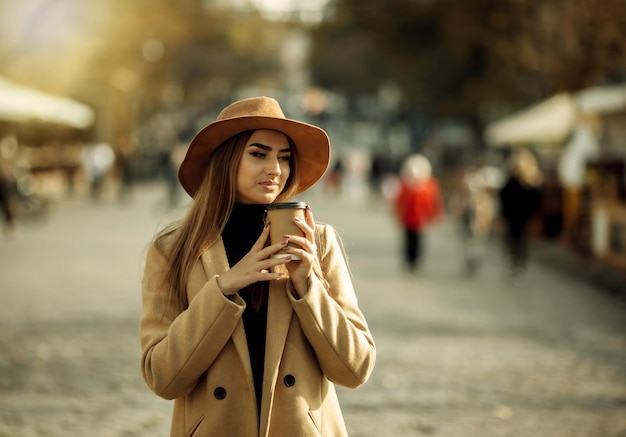Jonge stijlvolle vrouw gekleed in een jas en vilten hoed loopt door de stad en drinkt onderweg koffie. Herfst tijd