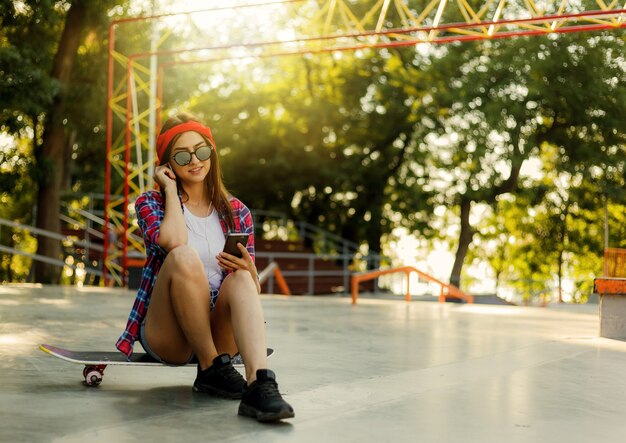 Jonge stijlvolle vrouw gekleed in jeugdkleren zit op een skateboard en luistert naar muziek op een koptelefoon in een skatepark. Zomertijd