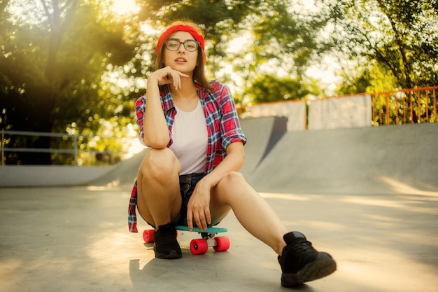 Jonge stijlvolle vrouw gekleed in jeugdkleren zit op een skateboard in een skatepark. Zomer, zonnige heldere dag