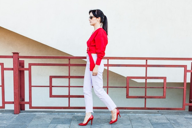 Jonge stijlvolle vrouw in een rode blouse en witte broek die in de stad loopt met schoenen met hoge hakken