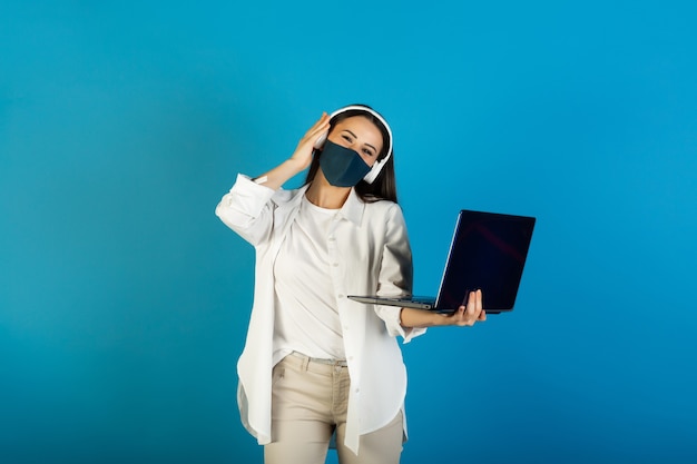 Jonge stijlvolle vrouw met gezichtsmasker en koptelefoon houdt laptopcomputer en luistert naar de muziek.