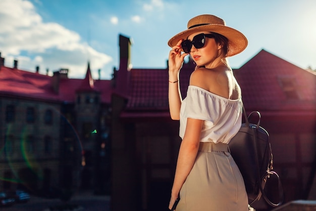 Jonge stijlvolle vrouw met hoed en zonnebril buitenshuis. Stijlvol meisje met rugzak en telefoon wandelen in Lviv met retro outfit