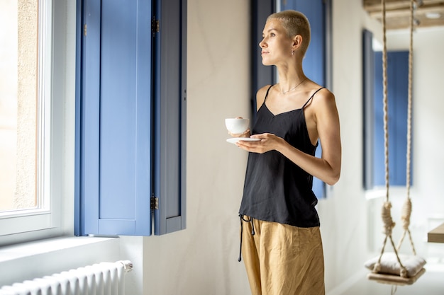 Jonge stijlvolle vrouw staat met een kopje koffie bij het raam in een modern, licht appartement. Comfortabel wonen en vrije tijd thuis