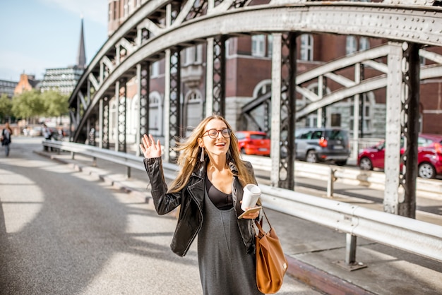 Jonge stijlvolle zakenvrouw met koffiekop die een taxi neemt op de oude ijzeren brug