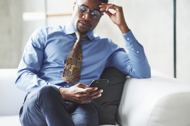 Jonge stijlvolle zwarte man in het pak en de bril die de telefoon vasthoudt en kijkt terwijl hij op de bank zit