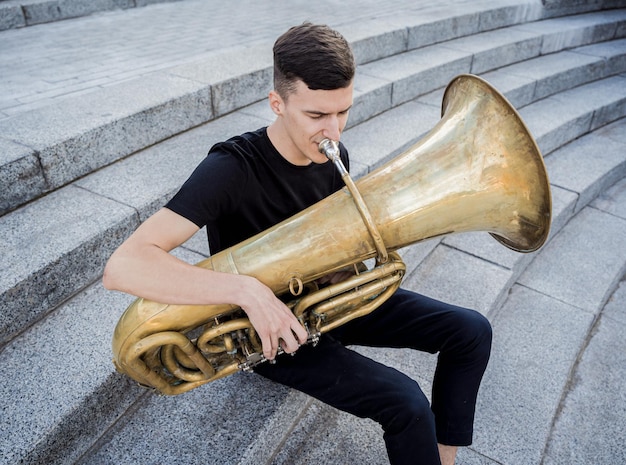 Jonge straatmuzikant speelt tuba zittend op granieten treden