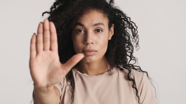 Jonge, strikte Afro-Amerikaanse vrouw die er zelfverzekerd uitziet en een stopgebaar toont op camera geïsoleerd op een witte achtergrond