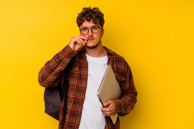 Jonge student blanke man met een laptop geïsoleerd op gele achtergrond met vingers op lippen die een geheim houden.