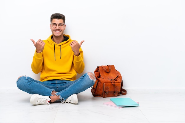 Jonge student blanke man zit op de vloer geïsoleerd op een witte achtergrond met duim omhoog gebaar en lachend