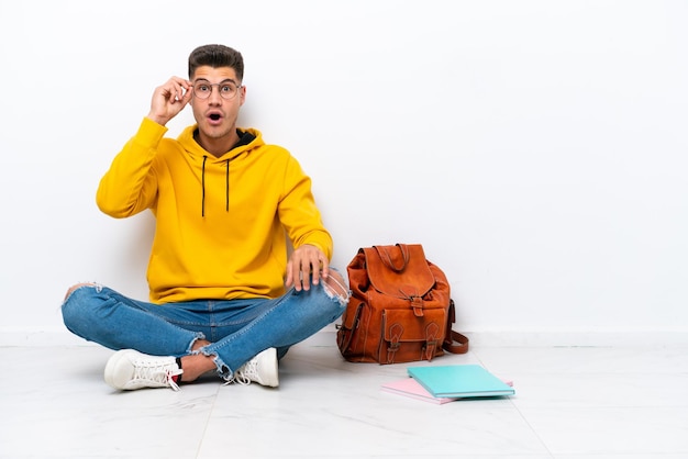 Jonge student blanke man zit op de vloer geïsoleerd op een witte achtergrond met een bril en verrast