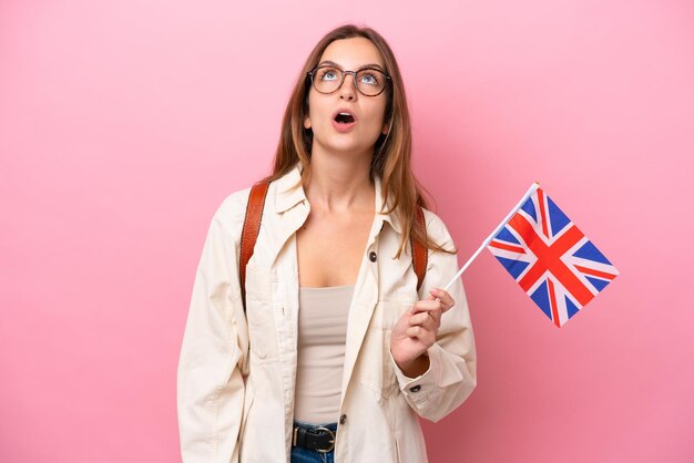 Jonge student blanke vrouw met een vlag van het Verenigd Koninkrijk geïsoleerd op roze achtergrond opzoeken en met verbaasde uitdrukking