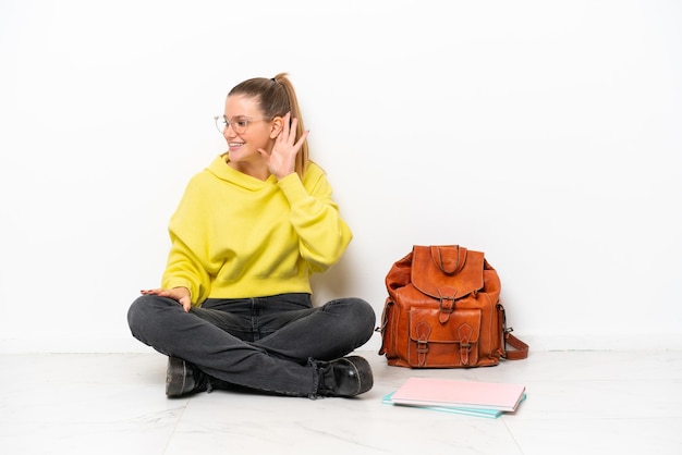 Jonge student blanke vrouw zittend op de vloer geïsoleerd op een witte achtergrond luisteren naar iets door hand op het oor te leggen