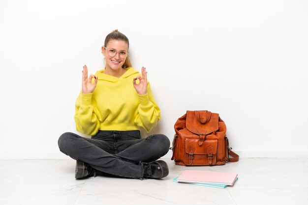 Jonge student blanke vrouw zittend op de vloer geïsoleerd op een witte achtergrond met een ok teken met vingers