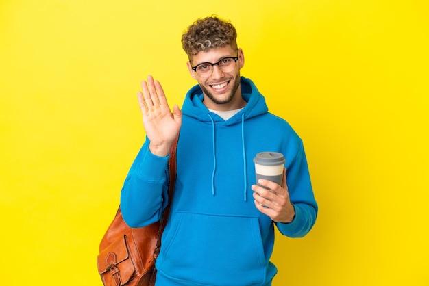 Jonge student blonde man geïsoleerd op gele achtergrond saluerend met de hand met gelukkige uitdrukking