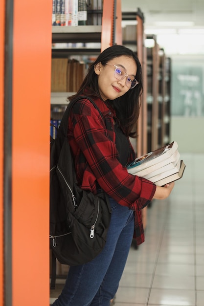 Jonge student die boeken vasthoudt en naar de camera kijkt terwijl hij in de buurt van de boekenkast in de bibliotheek staat