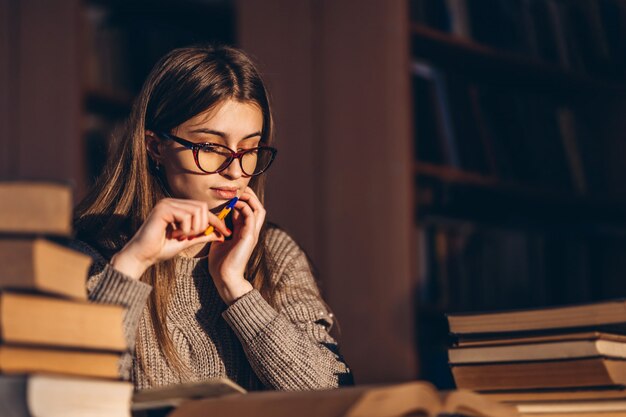 Jonge student die in glazen voor het examen voorbereidingen treft. Meisje in de avond zit aan een tafel in de bibliotheek met een stapel boeken