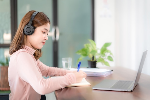 Jonge student draagt koptelefoon en schrijft op notebook terwijl hij thuis online studeert via laptop