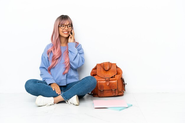 Jonge student gemengd ras vrouw met roze haar zittend op de vloer geïsoleerd op een witte achtergrond een gesprek voeren met de mobiele telefoon