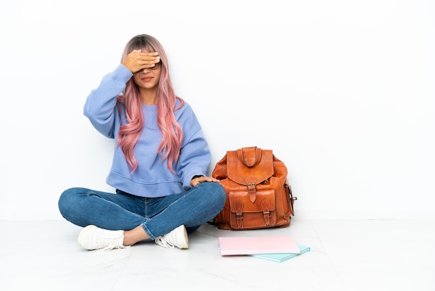 Jonge student gemengd ras vrouw met roze haren zittend op de vloer geïsoleerd op een witte achtergrond die ogen bedekt door handen. Wil je iets niet zien