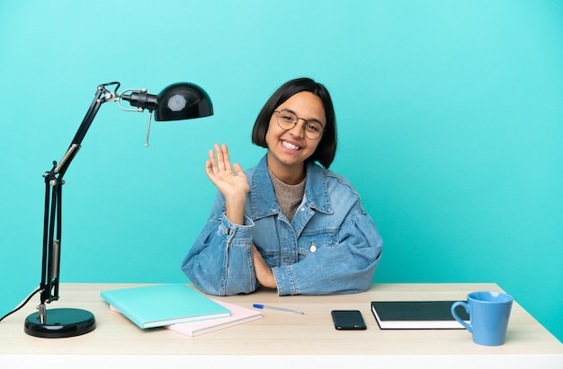 Jonge student gemengd ras vrouw studeren aan een tafel groeten met hand met gelukkige uitdrukking