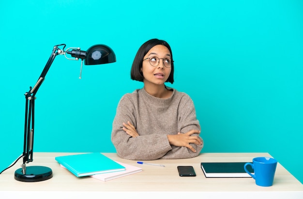 Jonge student gemengd ras vrouw studeren op een tafel terwijl ze glimlachend omhoog kijkt