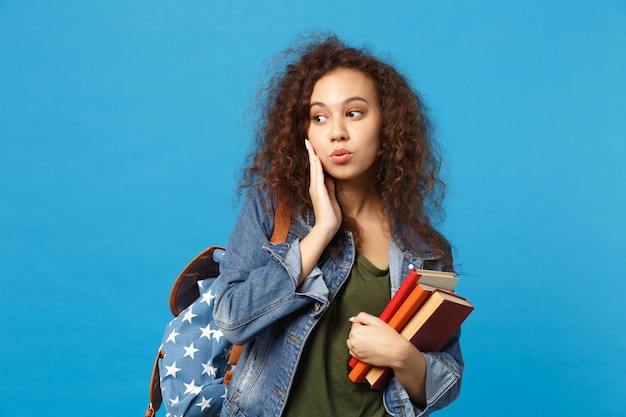 Jonge student in denim kleding en rugzak houdt boeken geïsoleerd op blauwe muur