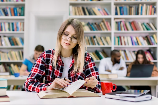 Jonge studente, casual geruit hemd en bril dragen, aan de tafel in de bibliotheek zitten en een boek lezen, voorbereiden op test of examen