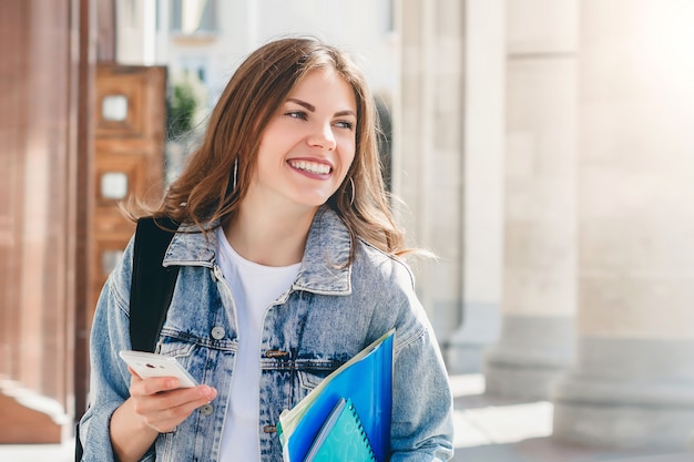 Jonge studente die tegen universiteit glimlacht. Leuke studente houdt mappen, laptops en mobiele telefoon in handen. Leren, onderwijs