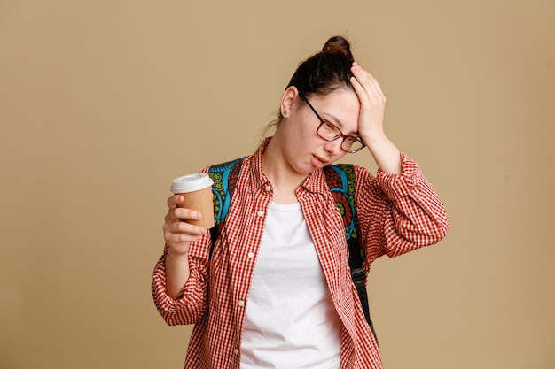 Foto jonge studente in vrijetijdskleding die een bril draagt met een rugzak die een koffiekopje vasthoudt en er verward uitziet, houdt de hand op haar hoofd voor een fout die over een bruine achtergrond staat