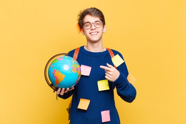 Jonge studentenjongen die vrolijk lacht, zich gelukkig voelt en naar de zijkant en naar boven wijst, een object in de kopieerruimte laat zien