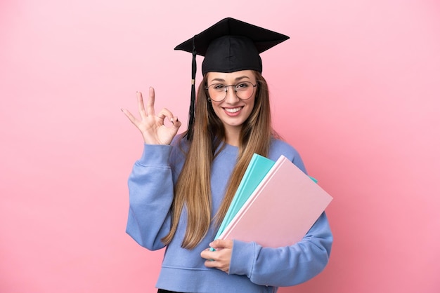 Jonge studentenvrouw die een gediplomeerde hoed draagt die op roze achtergrond wordt geïsoleerd die ok teken met vingers toont