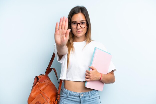 Jonge studentenvrouw die op blauwe achtergrond wordt geïsoleerd die stopgebaar maakt