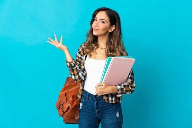 Jonge studentenvrouw die op blauwe muur wordt geïsoleerd die handen aan de kant uitbreidt om uit te nodigen om te komen