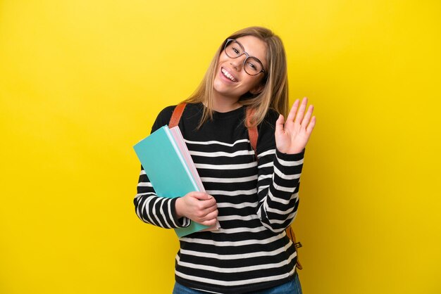 Jonge studentenvrouw die op gele achtergrondachtergrond wordt geïsoleerd die met hand met gelukkige uitdrukking salueert