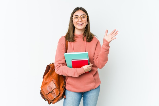 Jonge studentenvrouw die op witte bakcground wordt geïsoleerd die een aangename opgewekte verrassing ontvangt en handen opheft.