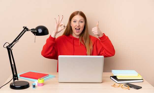 Jonge studentenvrouw op een werkplaats met laptop die ok teken en duim op gebaar tonen