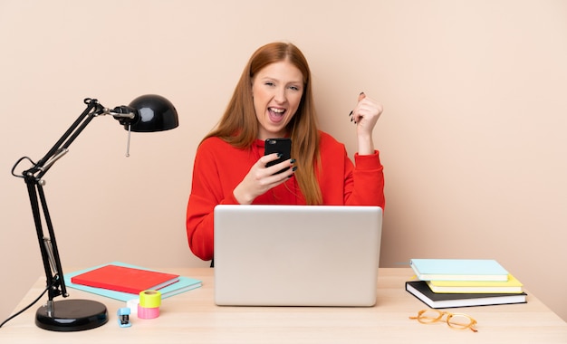 Jonge studentenvrouw op een werkplaats met laptop met telefoon in overwinningspositie