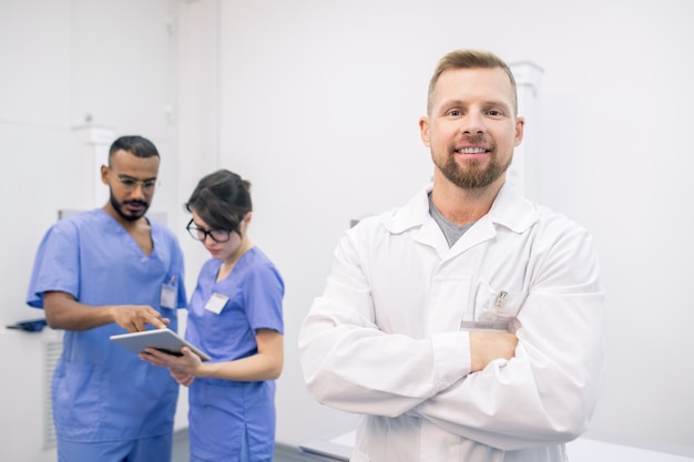 Jonge succesvolle bebaarde clinicus in whitecoat staan voor camera met zijn collega's die aan de achtergrond werken