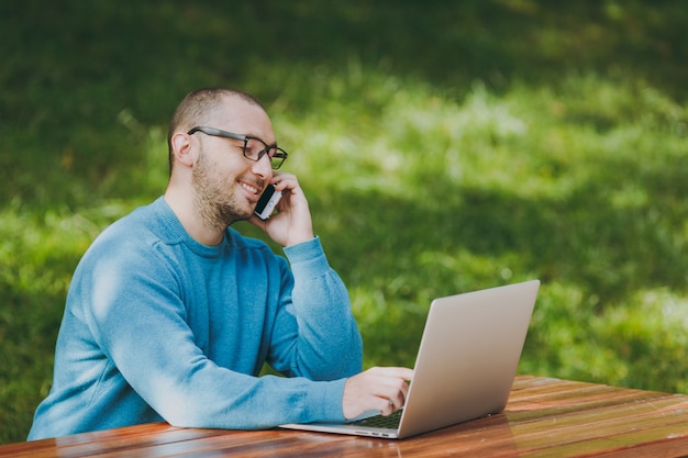 Jonge succesvolle lachende slimme man zakenman of student in casual blauw shirt, bril zittend aan tafel, praten op mobiele telefoon in stadspark met behulp van laptop, buitenshuis werken. Mobiel kantoorconcept.