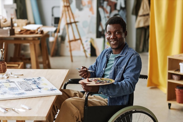 Jonge succesvolle mannelijke artiest met een handicap die palet vasthoudt met gemengde kleuren en penseel terwijl