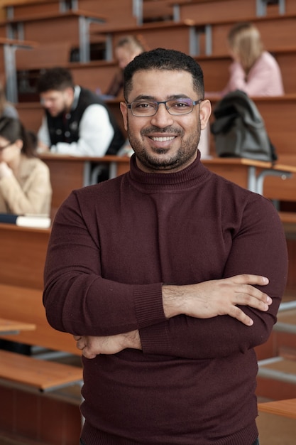 Jonge succesvolle mannelijke leraar die zijn armen kruist bij de borst terwijl hij voor de camera staat tegen studenten die bij een bureau in de collegezaal zitten