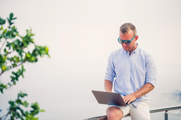 Jonge succesvolle mannelijke zakenman die met laptop op vakantie aan zee werkt Hij draagt een zonnebril, een shirt en een witte korte broek Werk buiten het kantoor freelancer