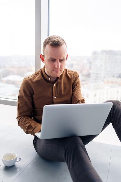 Jonge succesvolle mannelijke zakenman zit in de buurt van een groot raam met een laptop. Afstandswerk