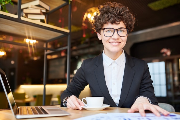 Jonge succesvolle onderneemster in formalwear en oogglazen die voor camera zitten terwijl het werken en het hebben van thee