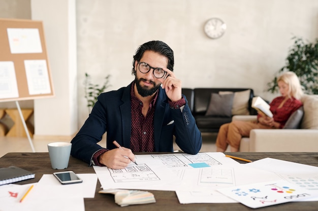 Jonge succesvolle ontwikkelaar van mobiele applicaties met een baard die een potlood op papier houdt met een schets terwijl hij aan een van de nieuwe items op kantoor werkt