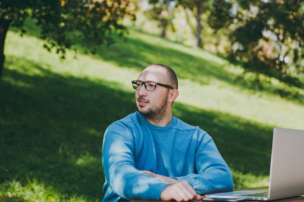 Jonge succesvolle slimme man zakenman of student in casual blauw shirt, bril zittend aan tafel met mobiele telefoon in stadspark met behulp van laptop, buitenshuis werken, opzij kijken. Mobiel kantoorconcept.